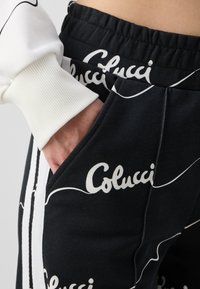 Pantalones de chándal negros con un patrón de texto blanco "Colucci", franjas laterales, cinturilla elástica y un bolsillo lateral. La tela parece suave y texturizada.
