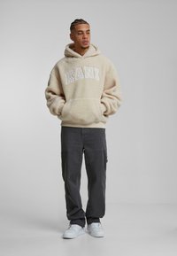 Felpa con cappuccio beige e soffice con la scritta "KANI" in lettere bianche, abbinata a pantaloni cargo grigi scuri e sneaker bianche.
