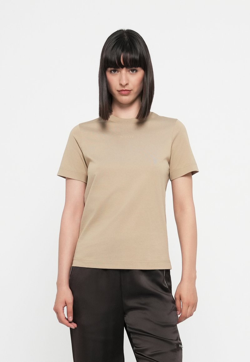 T-shirt beige a maniche corte in cotone. Presenta un colletto tondo e un discreto dettaglio del logo sulla parte anteriore. Texture liscia e omogenea.