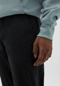 Světle modrá mikina s žebrovanými manžetami a černé kalhoty; detail ruky opírající se o nohavici kalhot, zobrazující texturu a detaily látky.