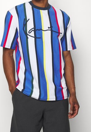 Mand iført en kortærmet t-shirt med lodrette striber i blå, hvid, rød, gul, grøn og marineblå, kombineret med mørke bukser.
