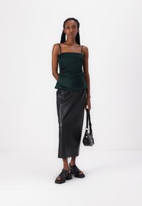 Green satin peplum top with thin straps, paired with a black faux leather midi skirt. Model holds a small black handbag and wears black platform sandals.