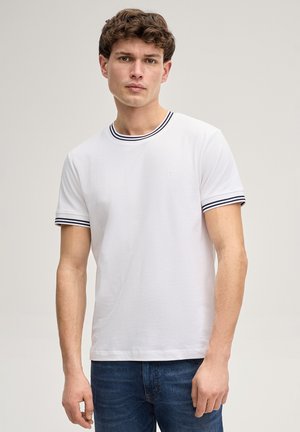 Junger Mann steht, trägt ein weißes kurzärmeliges T-Shirt mit navyblauem gestreiftem Kragen und Ärmelbündchen, kombiniert mit blauen Jeans.