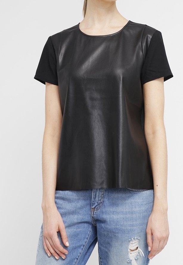 Femme portant un haut noir à manches courtes avec un panneau avant en similicuir et un jean bleu clair déchiré, debout devant un fond uni.