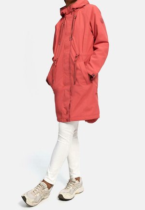 Personne portant un parka à capuche corail avec des cordons de serrage, un pantalon blanc et des baskets beige, debout jambes croisées sur un fond blanc.