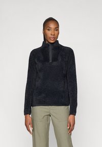 Sort fleece-pullover med høj krave og halv-lynlås foran. Blødt, struktureret materiale med lange ærmer, parret med olivengrønne bukser.