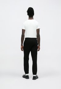 Sötét bőrű, rasztás hajú személy fehér pólót, fekete melegítőnadrágot, fehér zoknit és fekete cipőt visel, háttal áll egy fehér háttér előtt.