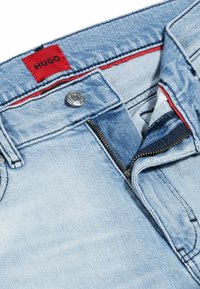 Jeans din denim bleu deschis cu un eticheta roșie HUGO, fermoar în față și închidere cu buton argintiu. Prezintă un material texturat și o căptușeală interioară de culoare bleu închis.