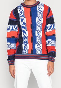 Gebreide sweater met een mix van rode, blauwe en witte patronen; geribbelde halslijn en manchetten; textiel met een structuur van geometrische ontwerpen.