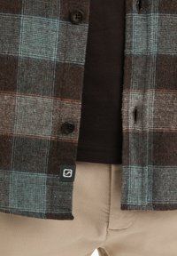 Geruite flanellen shirt in bruin, blauw en oranje tinten, met donkere knopen en een logotag. Daaronder een effen zwart shirt met beige broeken.