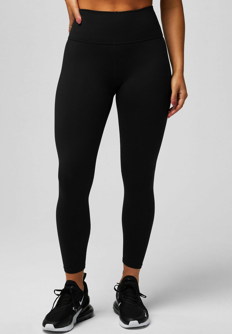 Schwarze hochtaillierte Leggings aus dehnbarem Stoff, mit einer glatten Textur und einer figurbetonten Passform, kombiniert mit schwarzen Sportschuhen.