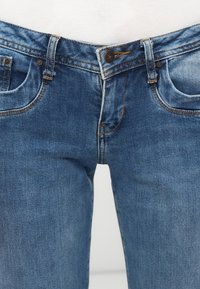 Priekša skatīta vidēja garuma zila denim auduma bikses ar pogu aizdari, jostas cilpām un priekšējām kabatām, kuras valkā persona ar baltu augšu.