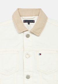 Biela denimová bunda s béžovým, štruktúrovaným kordurovým golierom. Má kovové gombíky a malú nášivku s logom v červenej, bielej a modrej farbe.