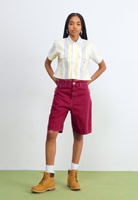Camicetta corta gialla chiaro con righe verticali blu e tasche, abbinata a pantaloni corti borgogna fino al ginocchio e stivali beige con lacci.
