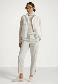 Colete acolchoado branco sobre um suéter de gola alta bege, combinado com calças brancas com aberturas laterais. Ténis brancos e óculos de sol amarelos oversized.