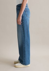 Hellblaue Denim-Weitleg-Jeans mit hoher Taille, ausgestattet mit zwei Gesäßtaschen und geradem Schnitt. Kombiniert mit hellen Sneakers.