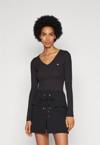 Top nero a maniche lunghe con scollatura a V e texture a costine, abbinato a una gonna nera con dettaglio di lace-up frontale e tasche laterali.