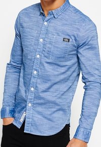 Jasnoniebieska, długosleevowa koszula zapinana na guziki, wykonana z materiału przypominającego dżins, z naszywką na lewej piersi i dwoma przednimi kieszeniami.