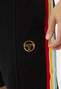Mustad mustad lühikesed püksid pehmest kangast, millel on kuldne ümmargune logo ja külje triibud punases, kollases ja teaalrohelises värvitoonis. Sile tekstuur, lihtne disain.