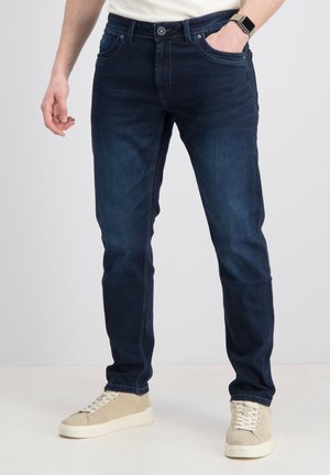 Man draagt donkerblauwe slim-fit jeans, beige sneakers en een wit shirt met een smartwatch aan de linkerpols, staande tegen een effen achtergrond.