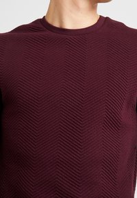 Uomo che indossa una maglietta a collo rotondo di colore viola scuro con un motivo chevron diagonale testurizzato sul tessuto.