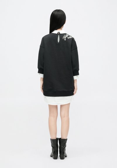 Μαύρη μπλούζα με φλοράλ κέντημα στην πλάτη, λευκό κολάρο ορατό από κάτω, συνδυασμένη με λευκή πουκάμισο και μαύρες μπότες μέχρι το γόνατο.