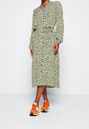 Fließendes grünes Kleid mit einem Leopardenmuster, langen Ärmeln und einem gebundenen Taillengürtel. Kombiniert mit orange-weißen Sneakers.