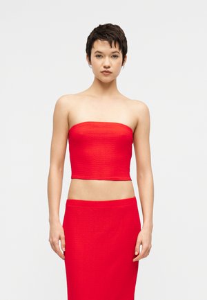 Woman with short dark hair wearing a red strapless crop top and matching high-waisted skirt, standing against a plain light background.