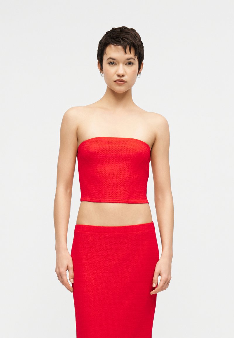 Γυναίκα με κοντά σκούρα μαλλιά που φοράει κόκκινο τοπ χωρίς τιράντες και φούστα ψηλόμεση που ταιριάζει, στέκεται μπροστά σε απλό φωτεινό φόντο.