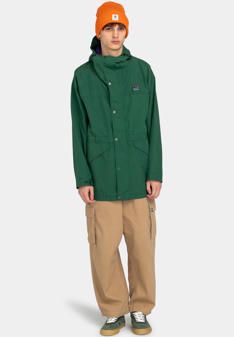 Giacca impermeabile verde con cappuccio, tasche frontali e bottoni a pressione, abbinata a pantaloni cargo beige e un berretto arancione. Sneakers verdi in esposizione.