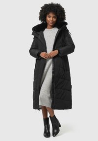 Manteau d'hiver noir long en duvet avec une capuche doublée de fourrure, design matelassé, fermeture éclair avant et poches latérales porté sur une robe en laine grise et des bottines noires.