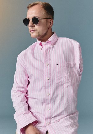Homme portant des lunettes de soleil et une chemise rose à rayures, posant avec la main droite reposant sur son genou sur un fond gris uni.