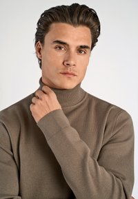 Maglione a collo alto marrone realizzato in morbido tessuto, con una silhouette aderente e polsini a coste. Texture liscia e design con colletto alto.