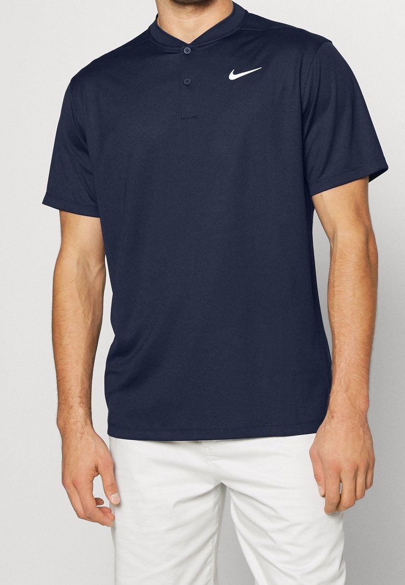 Homme portant un polo Nike bleu marine à manches courtes avec deux boutons et un pantalon blanc sur fond uni.