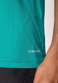 Gros plan d'un t-shirt de sport bleu sarcelle avec le texte "AEROREADY" sur le bas, porté par une personne avec un bras visible et un short noir.