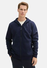 Marineblauwe rits-hoodie gemaakt van zachte stof, met een capuchon met trekkoord, zakken aan de voorkant, geribde mouwen en een logopatch op de mouw.