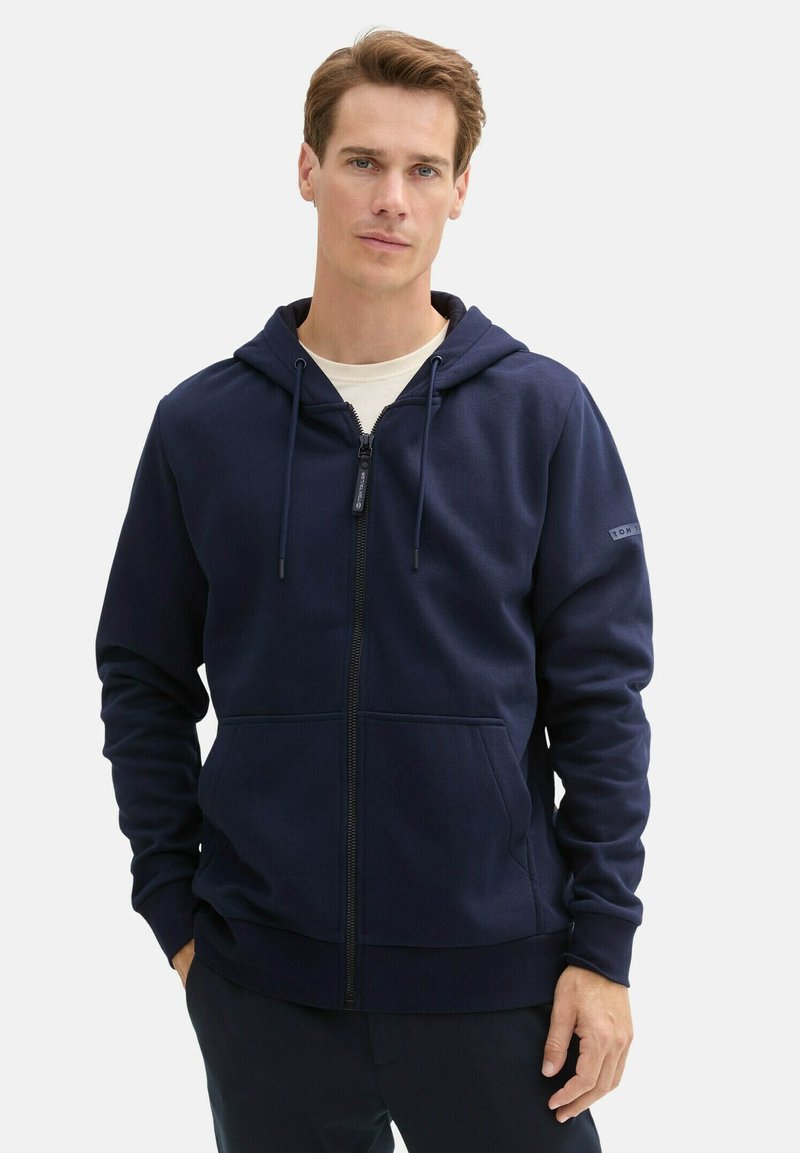 Marineblaue Zip-Hoodie aus weichem Stoff, ausgestattet mit einer Kapuze mit Kordelzug, Fronttaschen, ribbelten Bündchen und einem Logopatch am Ärmel.