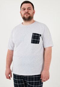 Siva kratka majica s kratkim rukavima s crnom četvrtastom džepom. Uparen s crnim pidžama hlačama s bijelim mrežastim uzorkom.