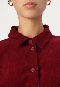 Bordeaux corduroy shirt met een klassieke kraag, twee knopen en een gestructureerde stof. De close-up benadrukt het materiaal en de kleur.