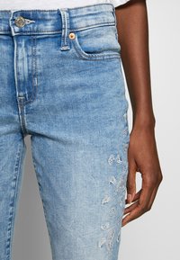 Jeans en denim bleu clair avec une coupe ajustée, ornés d'une broderie paisley argentée sur la cuisse gauche et des détails de poches standard.