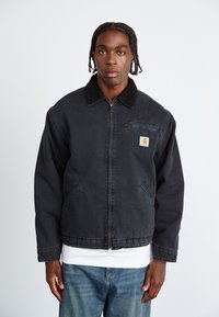 Mustmustest denim jakk, millel on pehme kaelus, ees lukk, küljetaskud ja rinna peal olev tasku logoga. Kantud valge särgi peal.