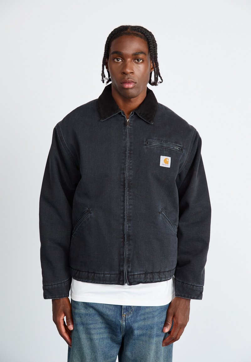 Mustmustest denim jakk, millel on pehme kaelus, ees lukk, küljetaskud ja rinna peal olev tasku logoga. Kantud valge särgi peal.