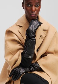 Black leather gloves with a sleek design and smooth texture, extending to the forearm. Worn with a camel-colored wool coat featuring a lapel.