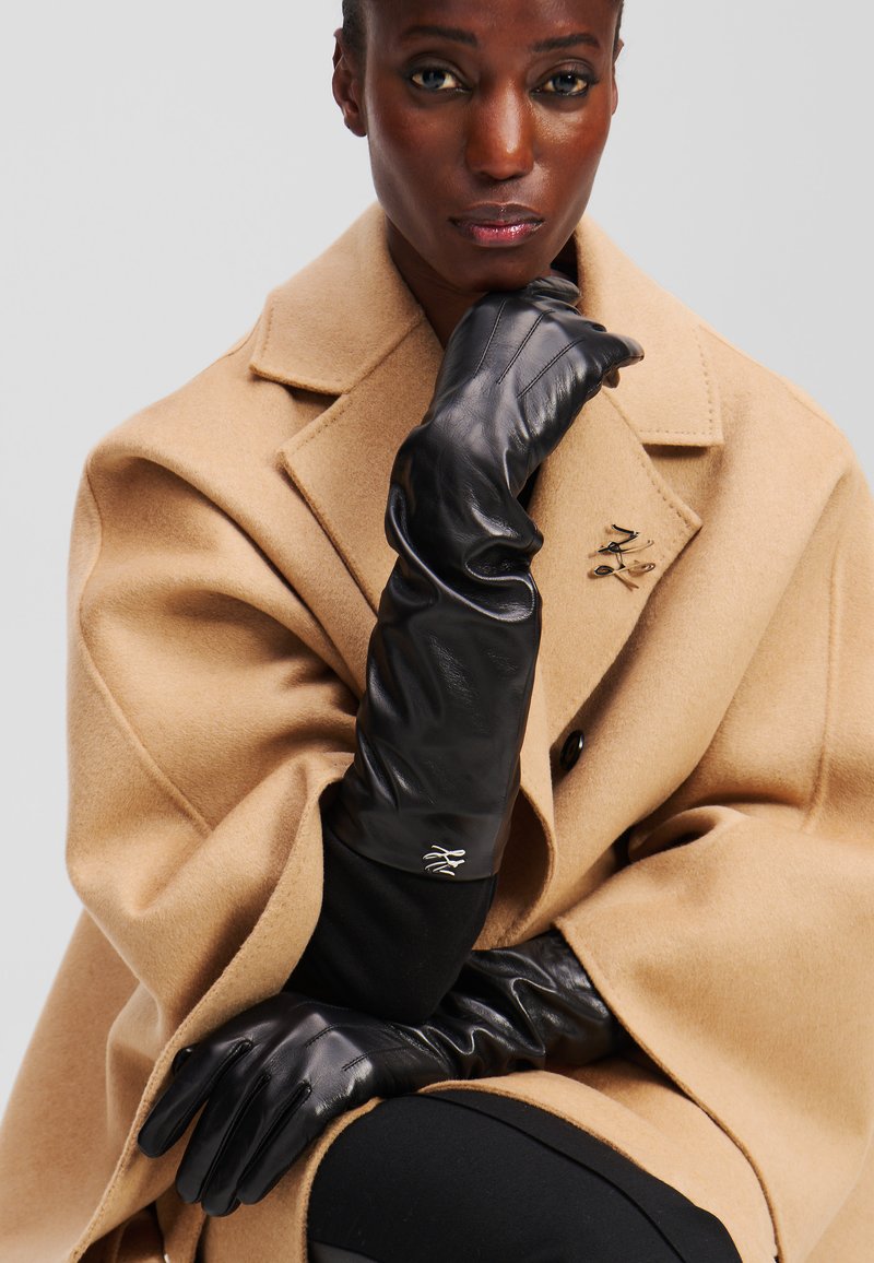 Black leather gloves with a sleek design and smooth texture, extending to the forearm. Worn with a camel-colored wool coat featuring a lapel.