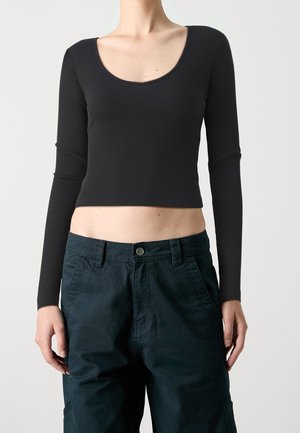 Persoon draagt nauwsluitende zwarte cropped top met lange mouwen en donkere, hooggetailleerde broek, staand tegen een eenvoudige lichte achtergrond.