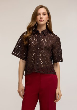 Vrouw met lang bruin haar die gouden oorbellen in de vorm van bladeren draagt, een donkerbruine kanten blouse met korte mouwen en knopen, en rode hoge taillebroeken.