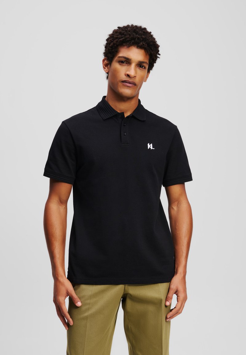 Melna polo krekls ar ribotu apkakli, īsām piedurknēm un mazu baltu logotipu kreisajā krūtī. Valkāts ar olīvzaļām biksēm.