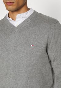 Maglione grigio a collo a V realizzato in tessuto a maglia. Presenta un piccolo logo ricamato in rosso, bianco e blu sul petto. Indossato sopra una camicia bianca.
