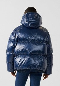 Giacca puffer blu navy con finitura lucida, dotata di cappuccio e design a righe orizzontali, indossata sopra un paio di jeans blu.