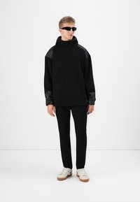 Felpa nera in cotone con colletto alto e pannelli strutturati sulle spalle, abbinata a pantaloni neri slim-fit e sneakers bianche.